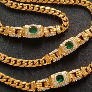 May include: A gold-toned jewelry set featuring a necklace and two bracelets. Each piece has a curb chain design with emerald-colored center stones surrounded by smaller, clear stones. The set is displayed on a dark surface.