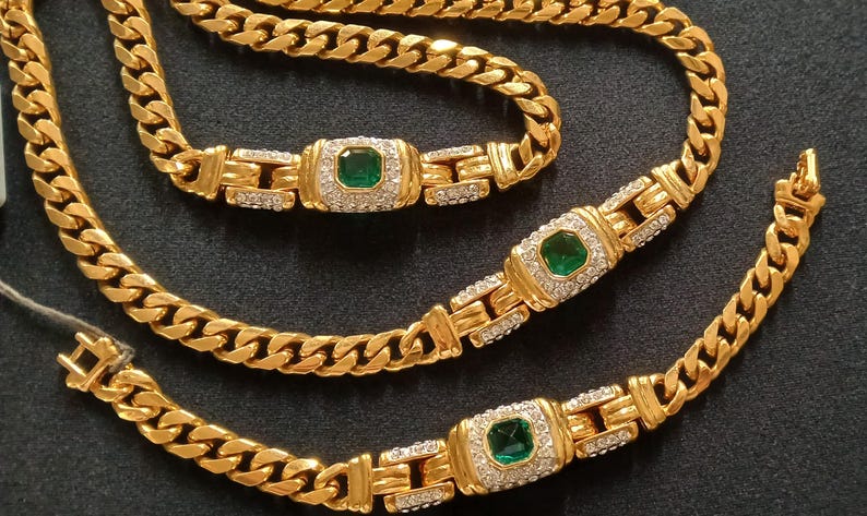 May include: A gold-toned jewelry set featuring a necklace and two bracelets. Each piece has a curb chain design with emerald-colored center stones surrounded by smaller, clear stones. The set is displayed on a dark surface.