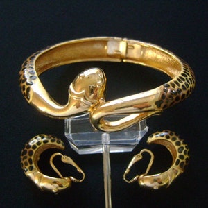 May include: A gold-tone snake-shaped bangle bracelet with black enamel detailing and matching hoop earrings. The jewelry set features a textured, spotted pattern.