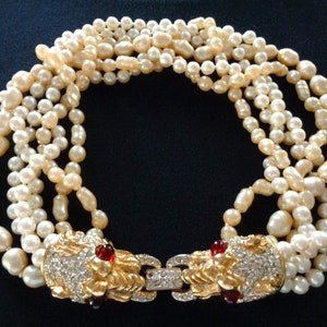 May include: A multi-strand pearl necklace with a gold clasp featuring two lion heads with red gemstone eyes and crystal accents.