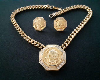 Puede incluir: Un collar de cadena dorada con un gran colgante octogonal que presenta un perfil detallado de la cara de una mujer. El colgante está adornado con pequeños diamantes de imitación transparentes. El collar también incluye un par de pendientes a juego con diseños similares.