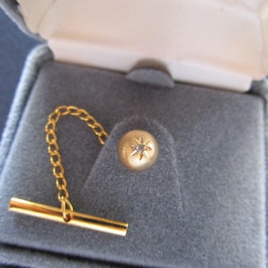 Op de afbeelding: Een gouden stropdasgesp met een klein diamantje in het midden. De stropdasgesp zit in een fluwelen doosje met een gouden ketting.