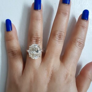 Op de afbeelding: Een hand met blauwe nagellak draagt een zilveren ring met een grote, heldere ovale edelsteen.
