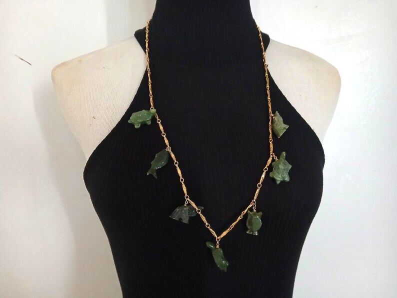 May include: A gold chain necklace with green jade turtle and fish charms. The necklace is on a black background.