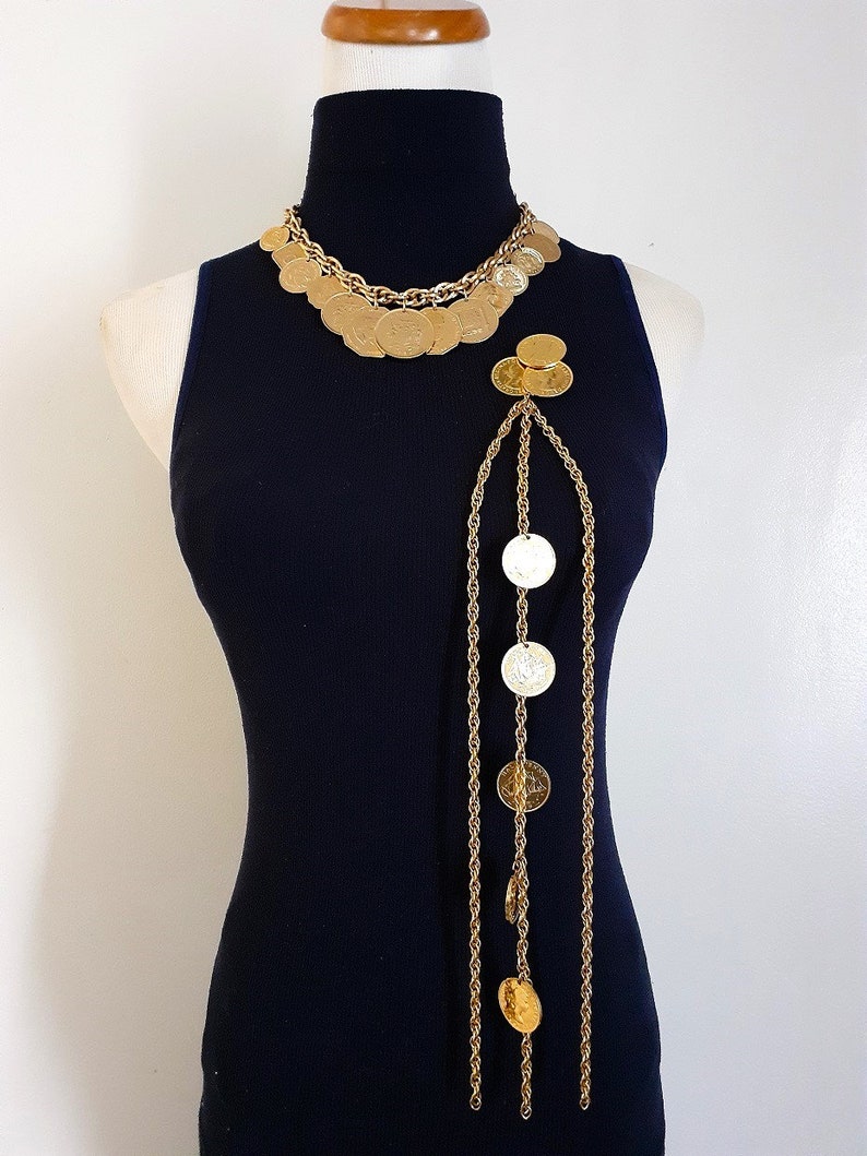 May include: A gold tone necklace with a long chain and multiple round coin-shaped charms. The necklace is layered with a shorter chain and more coin charms.