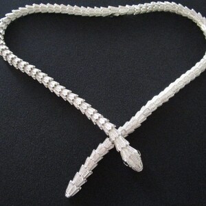 Op de afbeelding: Een zilveren slangenketting met twee slangenkoppen aan de uiteinden. De ketting ligt op een zwarte achtergrond.