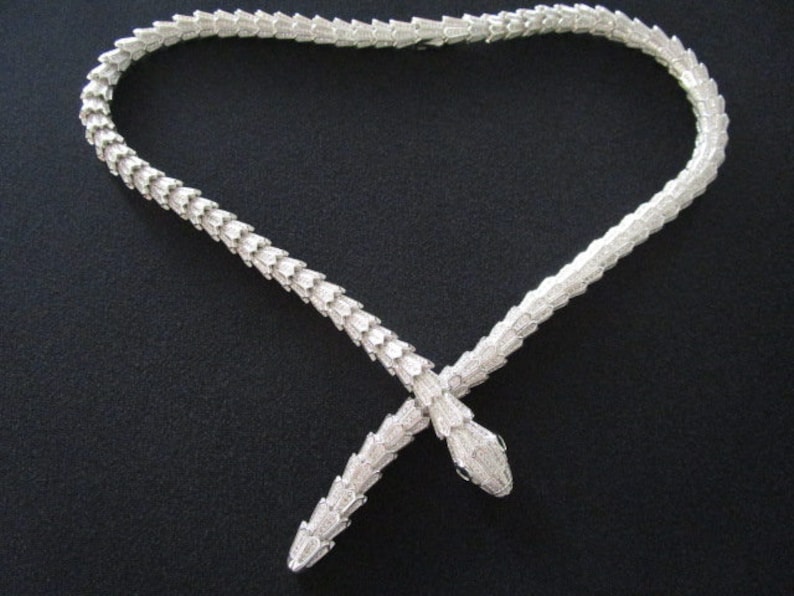 Op de afbeelding: Een zilveren slangenketting met twee slangenkoppen aan de uiteinden. De ketting ligt op een zwarte achtergrond.