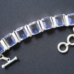 May include: A silver bracelet with a toggle clasp features a row of rectangular, faceted, light blue gemstones.