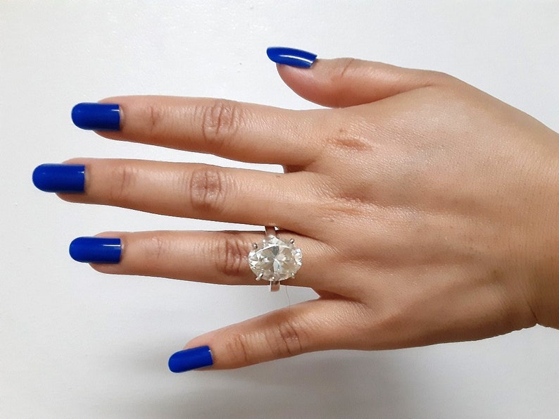 Op de afbeelding: Een hand met blauwe nagellak draagt een ring met een grote, heldere ovale edelsteen.