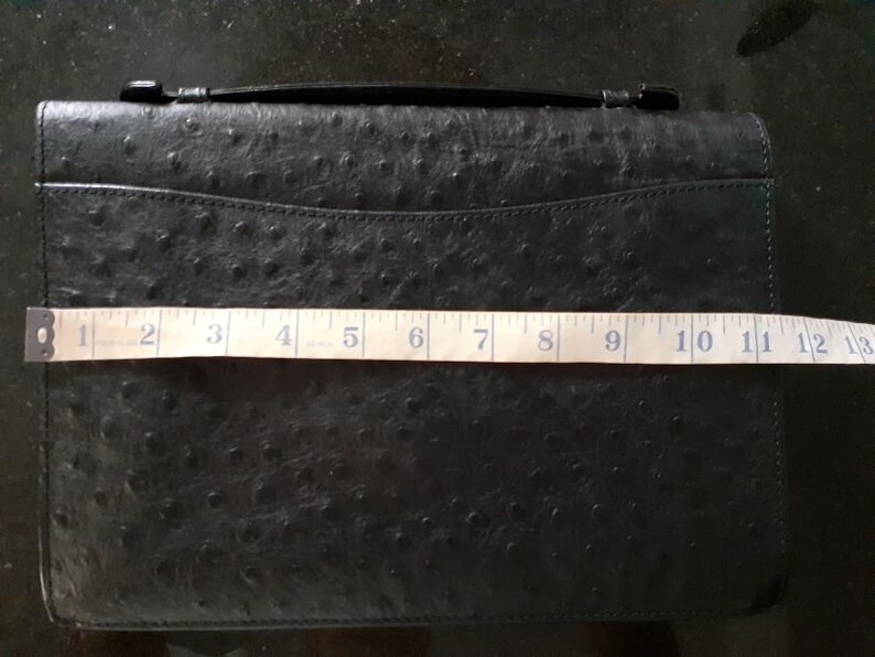 May include: Black ostrich leather clutch with a top handle and a zipper closure. The clutch measures 13 inches in length.