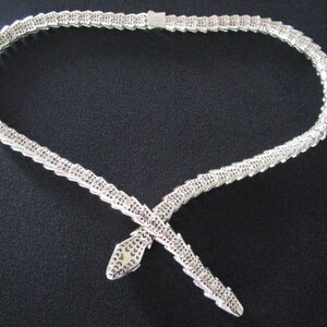Op de afbeelding: Een zilveren slangenketting met een gedetailleerd, gestructureerd ontwerp. De ketting is gemaakt van kleine, in elkaar grijpende schakels die een slangvorm vormen. De ketting is een statementstuk dat elke outfit een vleugje elegantie zou geven.