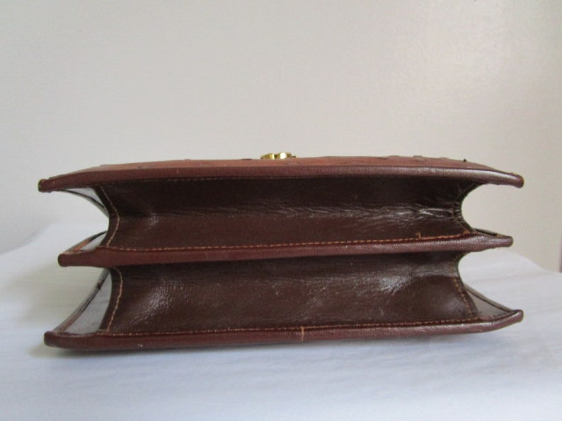 May include: A brown leather handbag with a flap closure and a gold-toned clasp. The bag has a structured shape and a double-layered design.