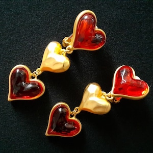 May include: A pair of gold-tone dangle earrings with red heart-shaped glass charms. The earrings have three hearts on each side, with the top heart being the largest.