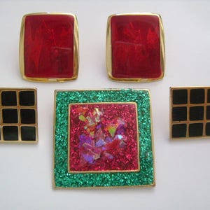 May include: Four gold-toned brooches with different designs. One is a red square with a gold frame, another is a black and gold grid pattern, another is a green and pink glitter design, and the last is a red square with a gold frame.