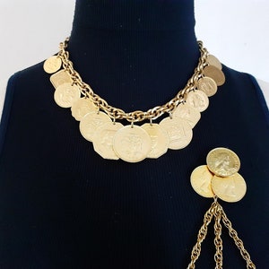 May include: A gold chain necklace with multiple gold coin charms. The necklace is on a black shirt. A separate gold chain with multiple gold coin charms is hanging from the bottom of the shirt.