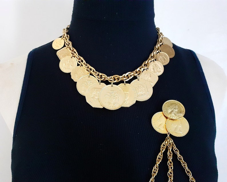 May include: A gold chain necklace with multiple gold coin charms. The necklace is on a black shirt. A separate gold chain with multiple gold coin charms is hanging from the bottom of the shirt.
