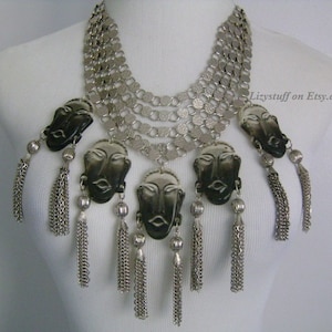 May include: A silver necklace with multiple chains and black face pendants. Each pendant has a silver tassel hanging from it. The necklace is on a white mannequin.