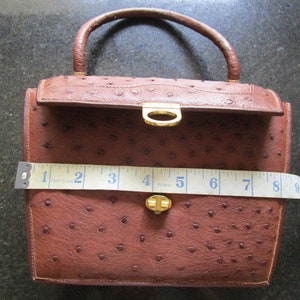 May include: A brown ostrich leather handbag with a gold-tone clasp and handle. The bag is approximately 7 inches wide.