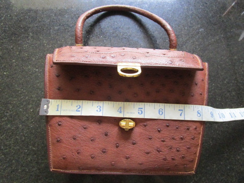 May include: A brown ostrich leather handbag with a gold-tone clasp and handle. The bag is approximately 7 inches wide.