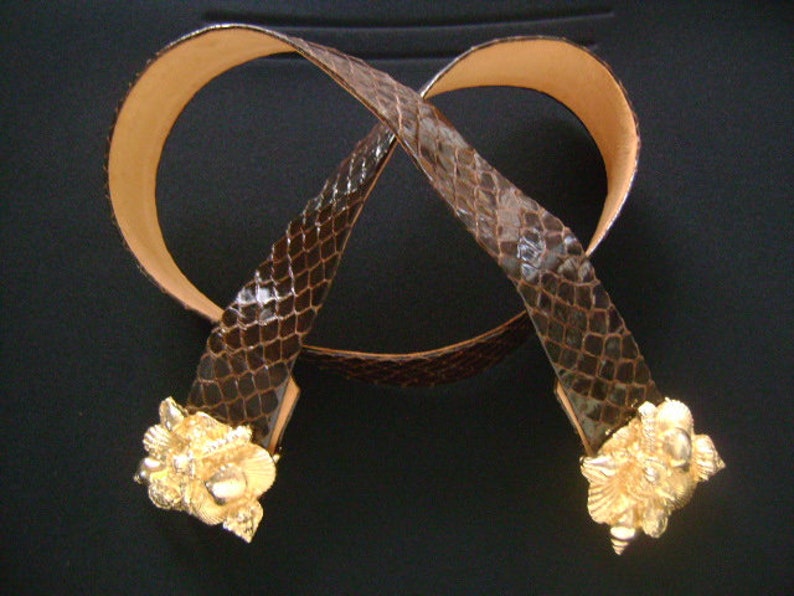 May include: A brown snakeskin belt with gold-toned decorative buckles. The belt is made of genuine leather and has a classic design.
