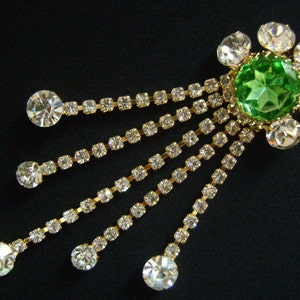 May include: A green rhinestone brooch with a flower design and dangling crystal chains. The brooch is made of gold-toned metal.
