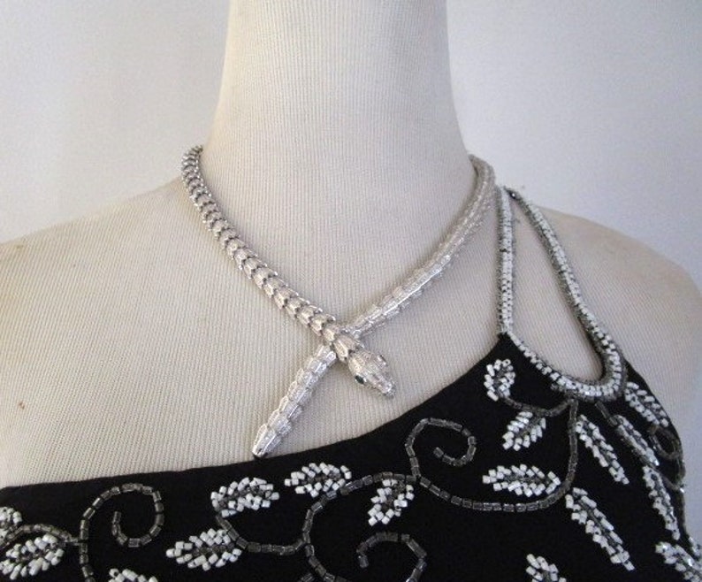 Op de afbeelding: Een zilveren slangenketting met een slangenkop-hanger. De ketting is op een mannequin die een zwart, met kralen bezet topje draagt.