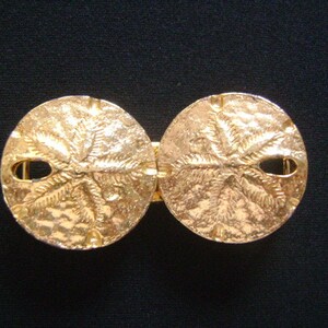 May include: A gold-tone belt buckle with a double-sided design featuring a sand dollar motif.