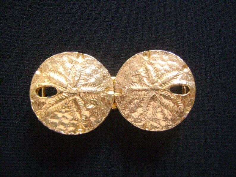 May include: A gold-tone belt buckle with a double-sided design featuring a sand dollar motif.