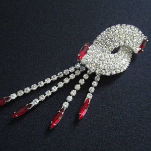 May include: A silver rhinestone brooch with a swirl design and red rhinestone accents. The brooch has a dangling chain with red rhinestones.