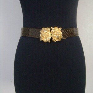 May include: A brown snakeskin belt with a gold-toned double buckle. The buckle is ornate and features a floral design.