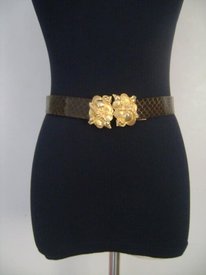 May include: A brown snakeskin belt with a gold-toned double buckle. The buckle is ornate and features a floral design.