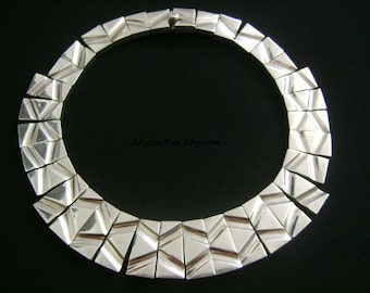 May include: A silver geometric necklace made of interlocking triangular pieces. The necklace is a statement piece with a modern design.