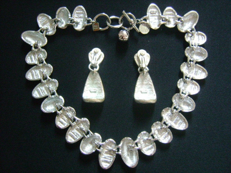 Puede incluir: Un collar de plata con una cadena de eslabones ovalados conectados y pendientes colgantes de plata a juego. El collar tiene un cierre de plata.