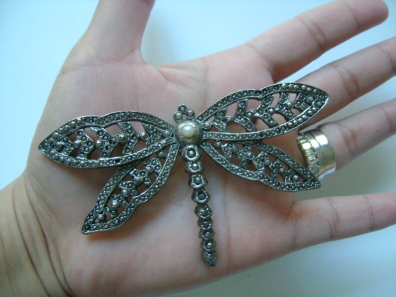 May include: A silver dragonfly brooch with a pearl accent. The dragonfly is made of intricate metalwork and has a delicate design.