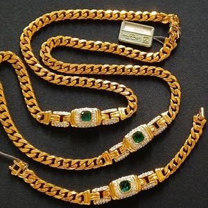 May include: A gold-colored necklace and bracelet set. The necklace and bracelet feature a curb chain design with emerald-colored stones and small clear stones. The set is displayed on a black surface.