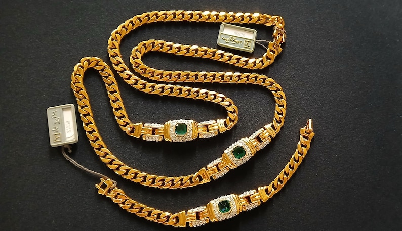 May include: A gold-colored necklace and bracelet set. The necklace and bracelet feature a curb chain design with emerald-colored stones and small clear stones. The set is displayed on a black surface.