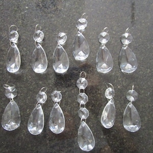 Op de afbeelding: Een verzameling van 12 heldere kristallen traanvormige kroonluchterprisma's met verschillende lengtes. Sommige prisma's hebben een enkele traanvorm, terwijl andere meerdere tranen hebben die aan elkaar zijn verbonden.