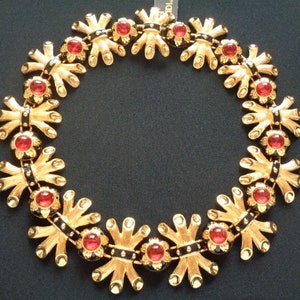 May include: A gold-toned necklace with a repeating floral design. Each flower has a red center stone and is connected by black enamel links. The necklace is displayed on a dark background.
