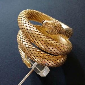 May include: A gold-colored snake bracelet coiled around itself. The snake's head is detailed with engraved patterns, and the body is made of small, connected scales. The bracelet is displayed on a clear stand against a dark background.