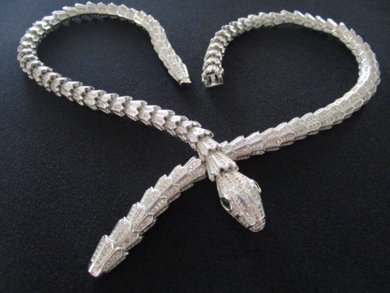 Op de afbeelding: Een zilveren slangenketting met een gedetailleerd, gestructureerd ontwerp. De ketting is gemaakt van metaal en heeft een slangenk kop met een groene stenen oog.
