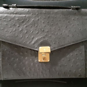 May include: A black leather briefcase with a textured ostrich skin pattern. The briefcase has a gold-toned lock and clasp.