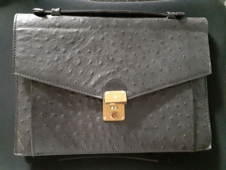 May include: A black leather briefcase with a textured ostrich skin pattern. The briefcase has a gold-toned lock and clasp.