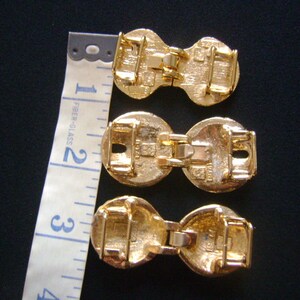 May include: Three gold-toned metal belt buckles with a decorative design. The buckles are connected by a metal bar.
