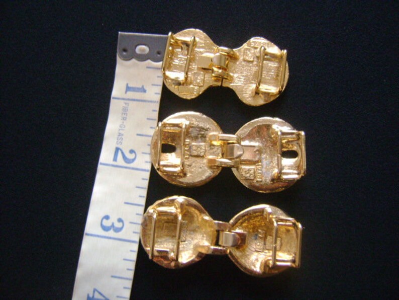 May include: Three gold-toned metal belt buckles with a decorative design. The buckles are connected by a metal bar.