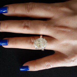 Op de afbeelding: Een grote, heldere ovale diamanten ring gezet in een zilveren band. De ring zit aan een vinger.