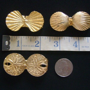May include: Two pairs of gold-tone metal clasps. One pair is shaped like seashells and the other pair is shaped like sand dollars. The clasps are shown next to a ruler with inches marked.