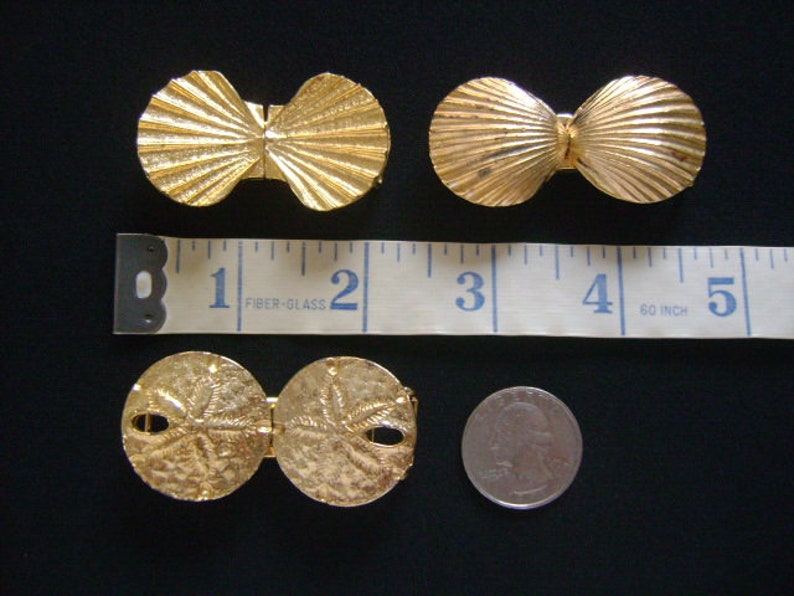May include: Two pairs of gold-tone metal clasps. One pair is shaped like seashells and the other pair is shaped like sand dollars. The clasps are shown next to a ruler with inches marked.