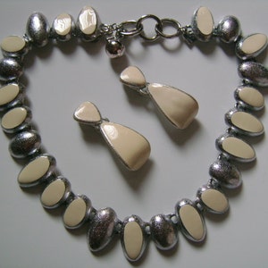 Puede incluir: Un collar de plata con una cadena y colgantes ovalados de color crema. Pendientes a juego de color crema con detalles plateados.