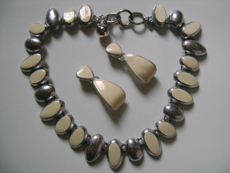 Puede incluir: Un collar de plata con una cadena y colgantes ovalados de color crema. Pendientes a juego de color crema con detalles plateados.