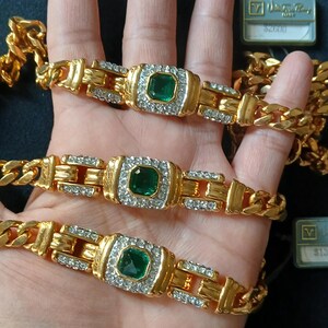 May include: Gold-toned bracelets with emerald-colored center stones and clear accent stones. The bracelets are displayed on a hand, with a gold chain visible. The bracelets have a rectangular design.
